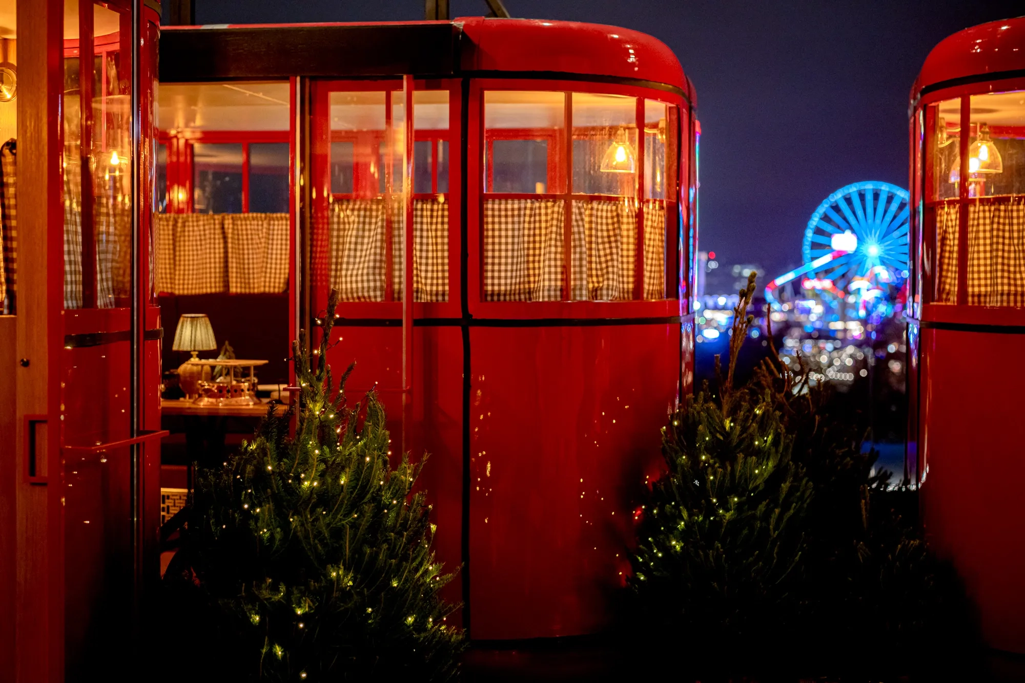 Red ski gondolas decorated with warm lights and small Christmas trees on a rooftop, with a lit Ferris wheel in the distance