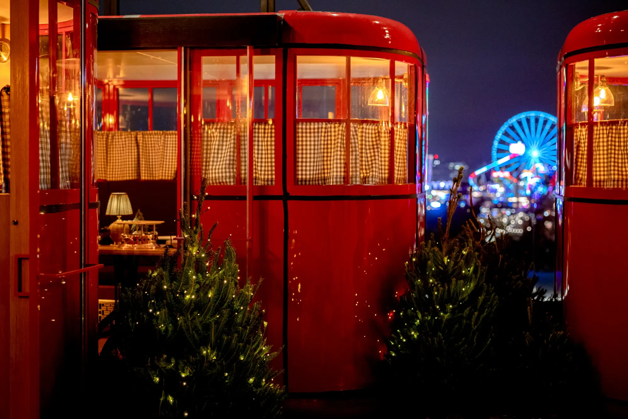Red ski gondolas decorated with warm lights and small Christmas trees on a rooftop, with a lit Ferris wheel in the distance