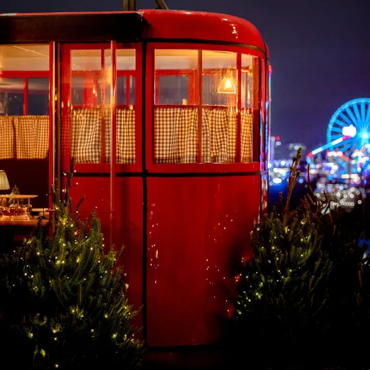 Red ski gondolas decorated with warm lights and small Christmas trees on a rooftop, with a lit Ferris wheel in the distance