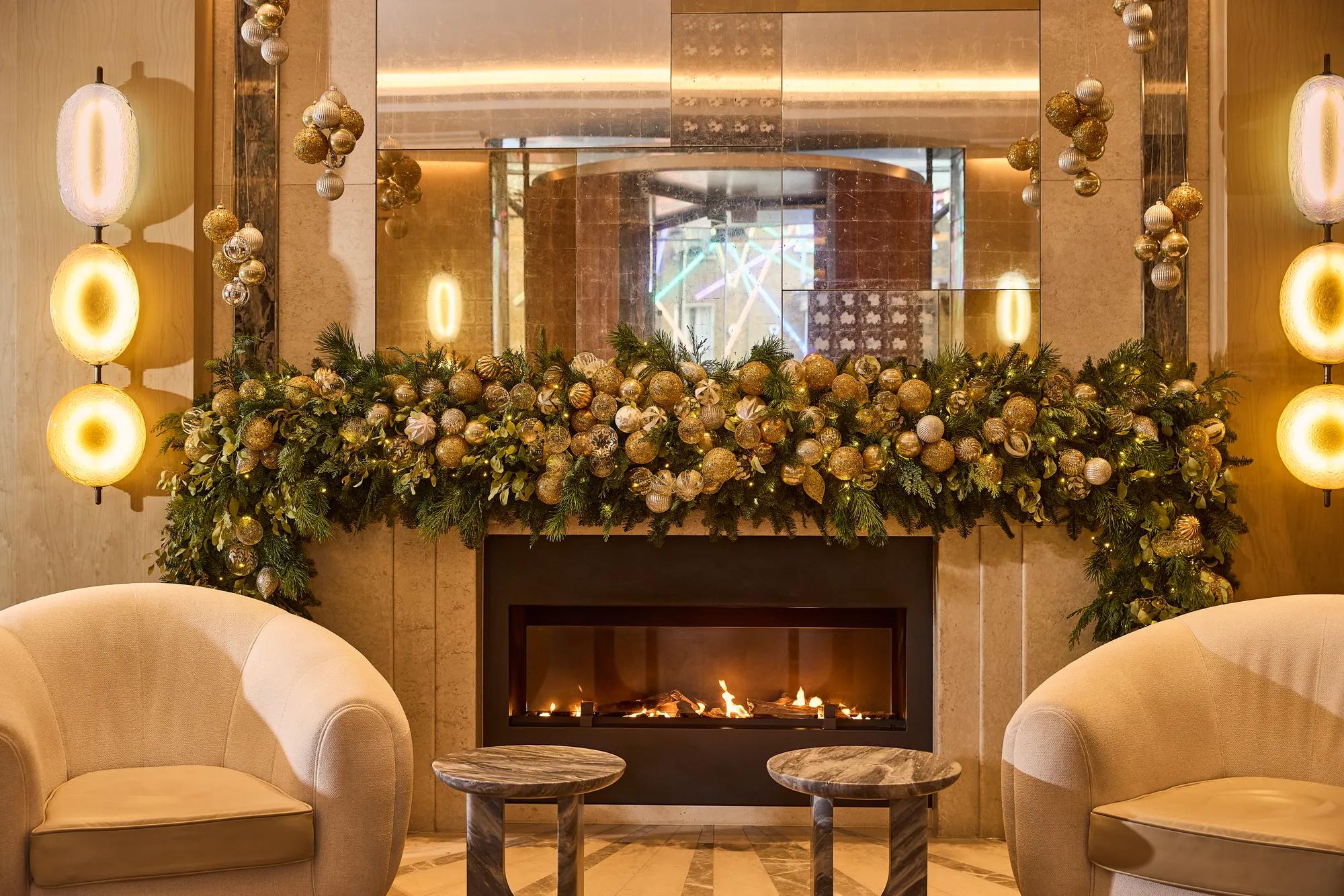 Festively decorated fireplace with garland, ornaments, armchairs and glowing lights in a luxurious lounge setting.
