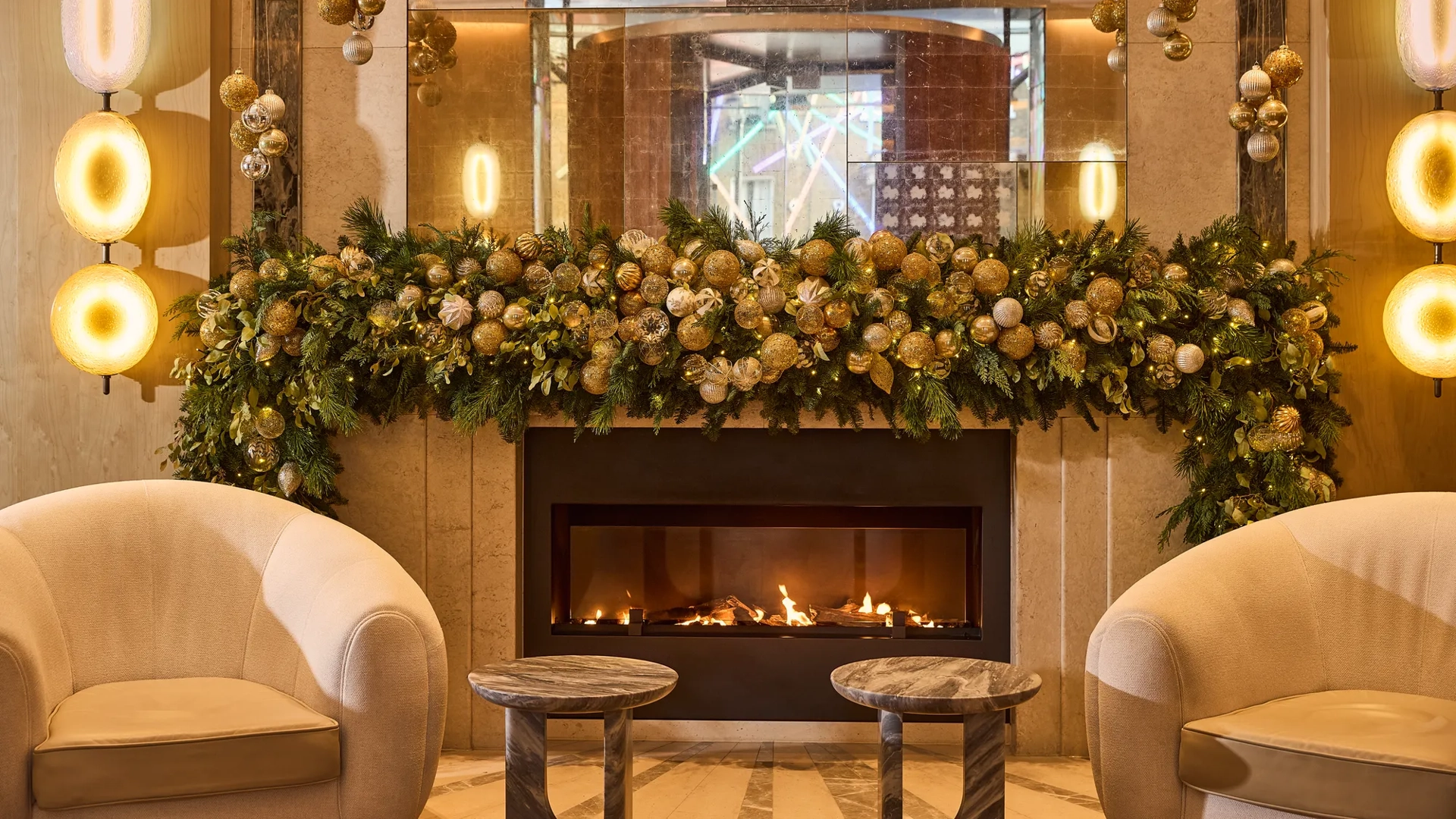 Festively decorated fireplace with garland, ornaments, armchairs and glowing lights in a luxurious lounge setting.