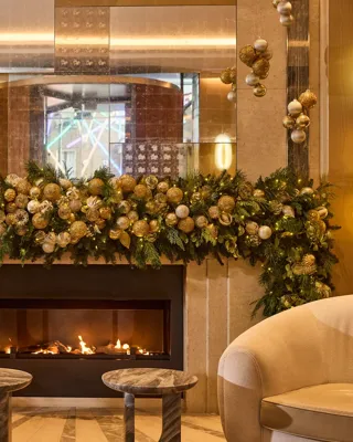 Festively decorated fireplace with garland, ornaments, armchairs and glowing lights in a luxurious lounge setting.