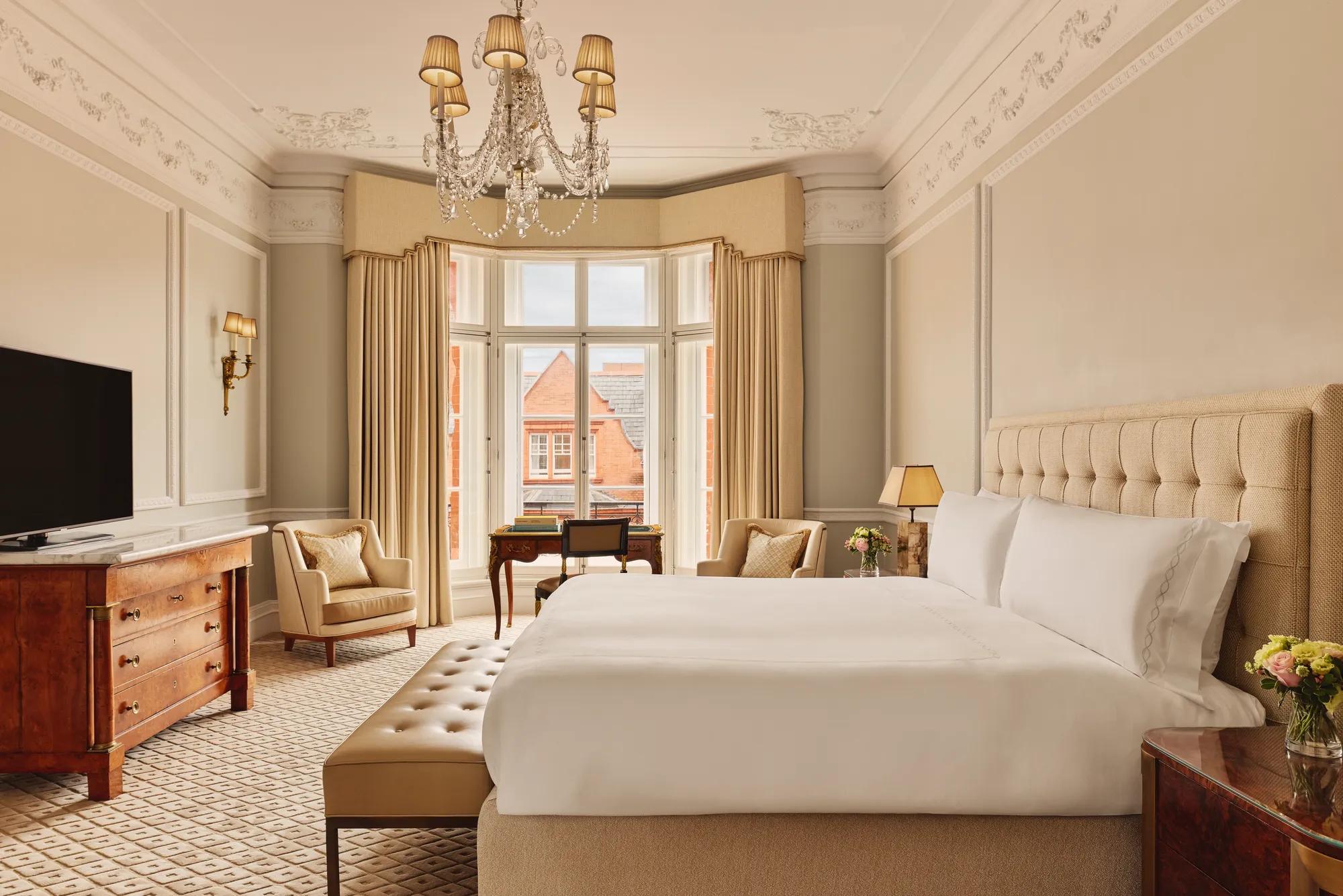 Elegant hotel bedroom with a large bed dressed in white linens, beige upholstered headboard, chandelier, and bay window with views of red-brick rooftops, featuring classic wooden furniture and neutral décor.