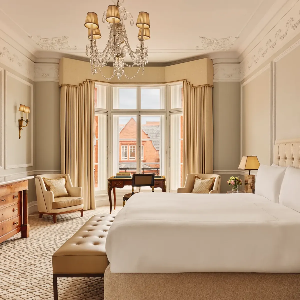 Elegant hotel bedroom with a large bed dressed in white linens, beige upholstered headboard, chandelier, and bay window with views of red-brick rooftops, featuring classic wooden furniture and neutral décor.