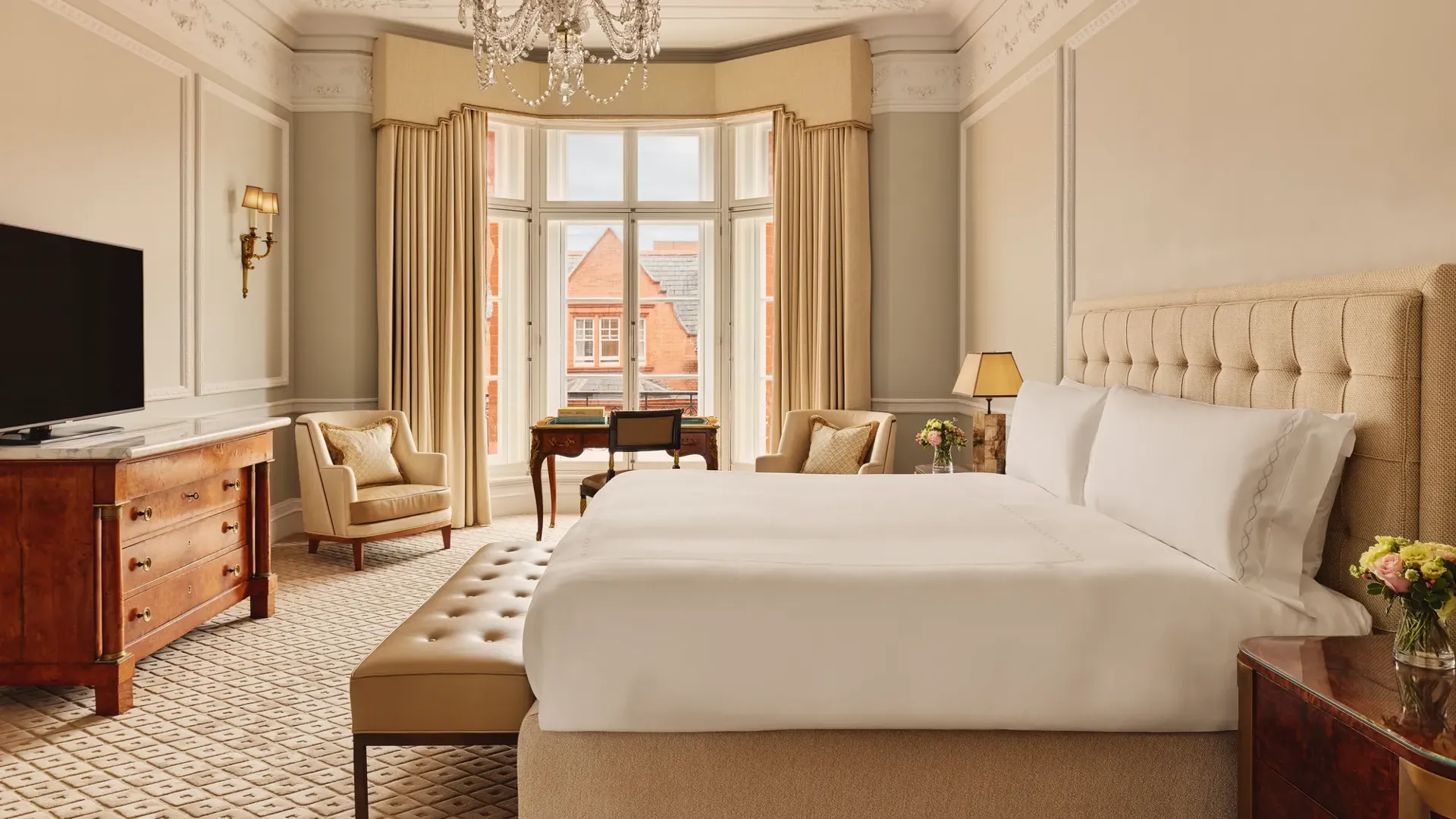 Elegant hotel bedroom with a large bed dressed in white linens, beige upholstered headboard, chandelier, and bay window with views of red-brick rooftops, featuring classic wooden furniture and neutral décor.