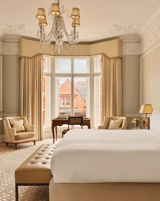 Elegant hotel bedroom with a large bed dressed in white linens, beige upholstered headboard, chandelier, and bay window with views of red-brick rooftops, featuring classic wooden furniture and neutral décor.