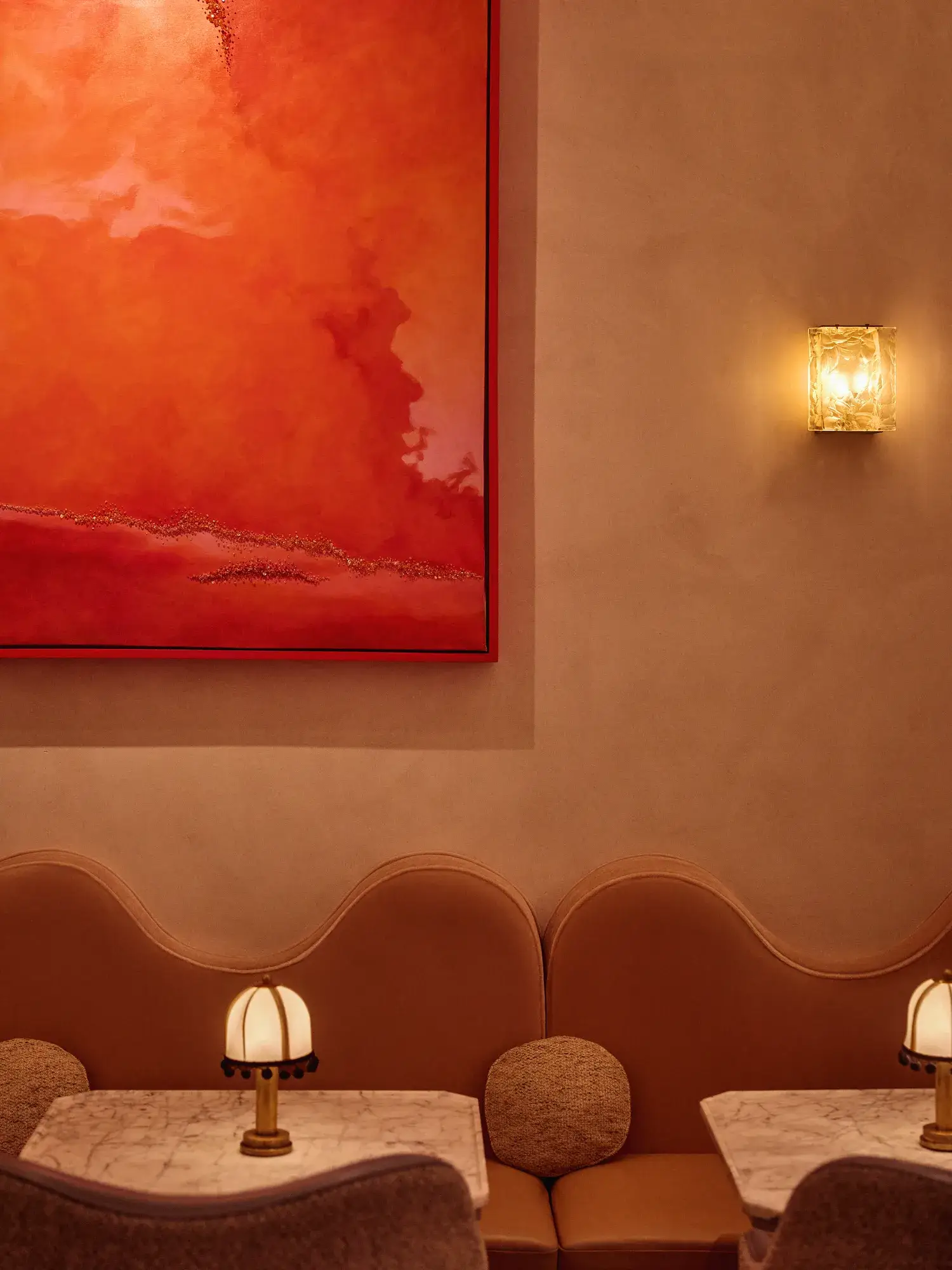 Softly lit lounge seating with curved tan banquette, marble tables, small lamps, and a large red abstract painting above.