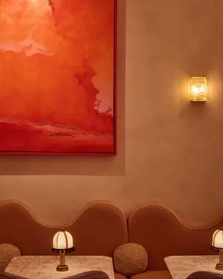 Softly lit lounge seating with curved tan banquette, marble tables, small lamps, and a large red abstract painting above.