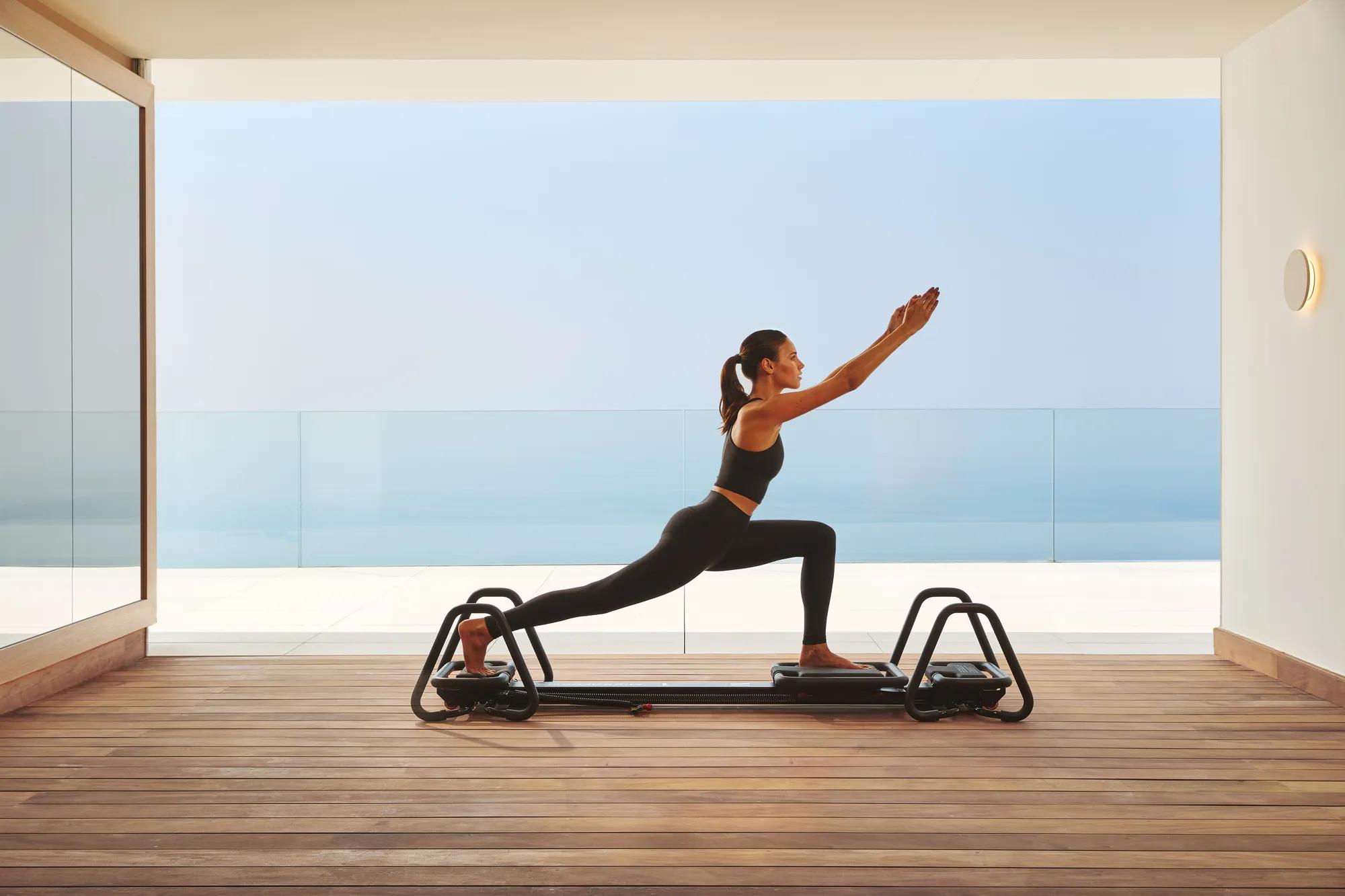 Woman practicing Pilates on a reformer in a modern studio with ocean view through glass walls.