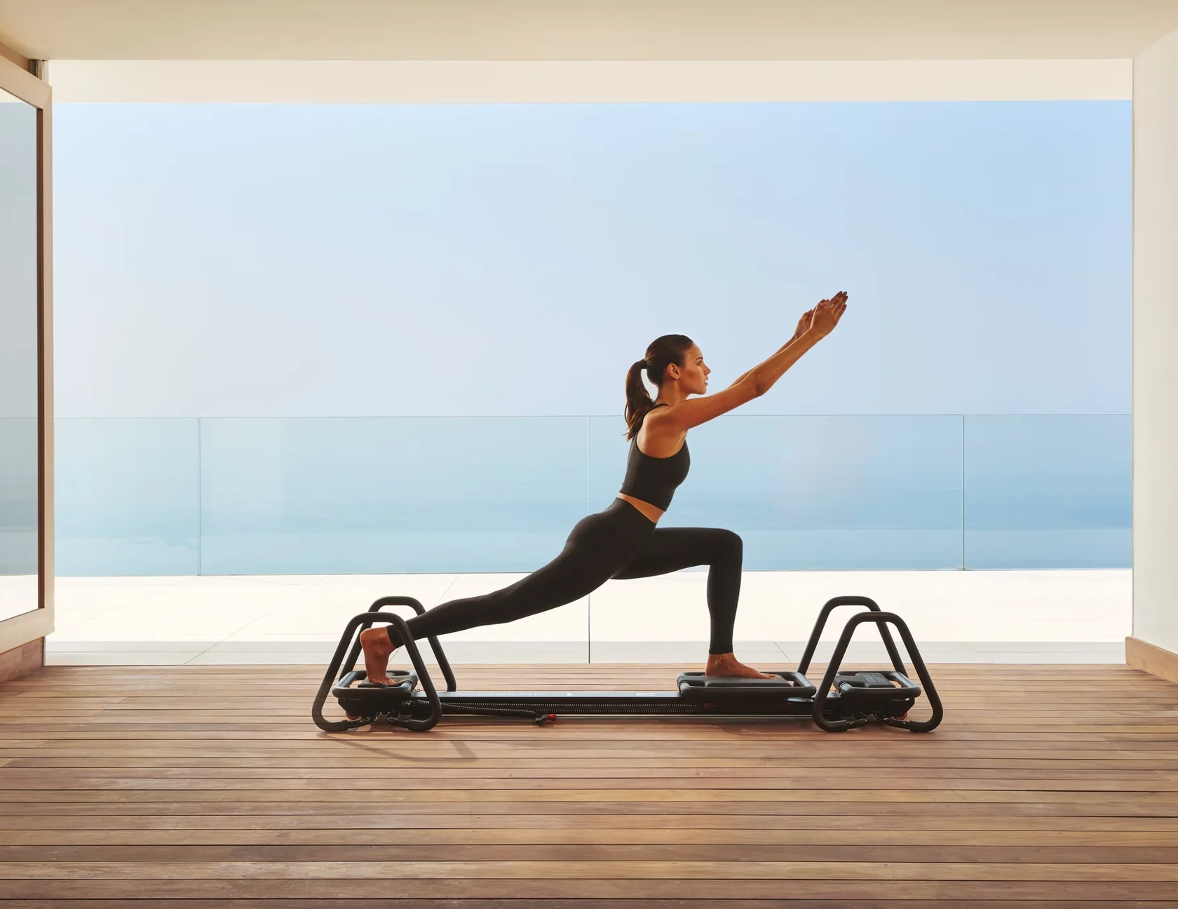 Woman practicing Pilates on a reformer in a modern studio with ocean view through glass walls.