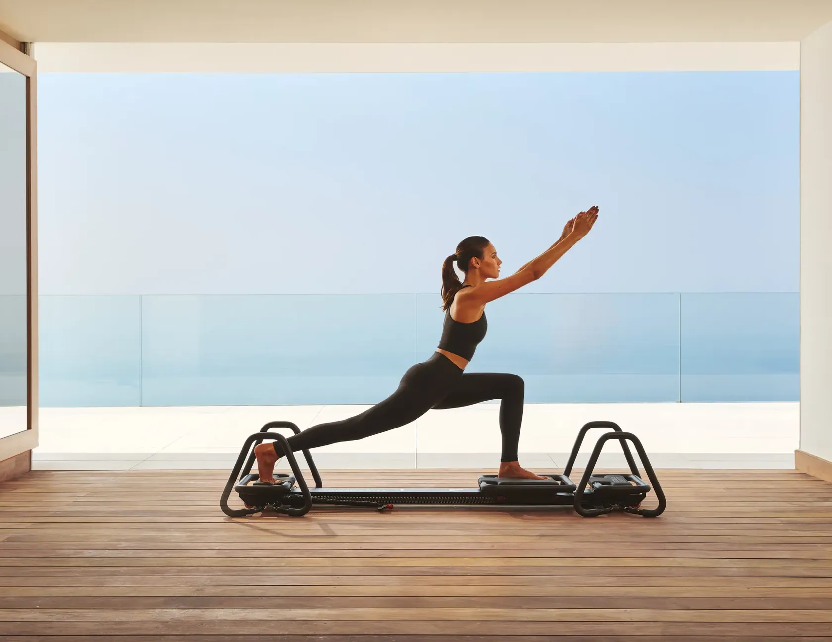 Woman practicing Pilates on a reformer in a modern studio with ocean view through glass walls.