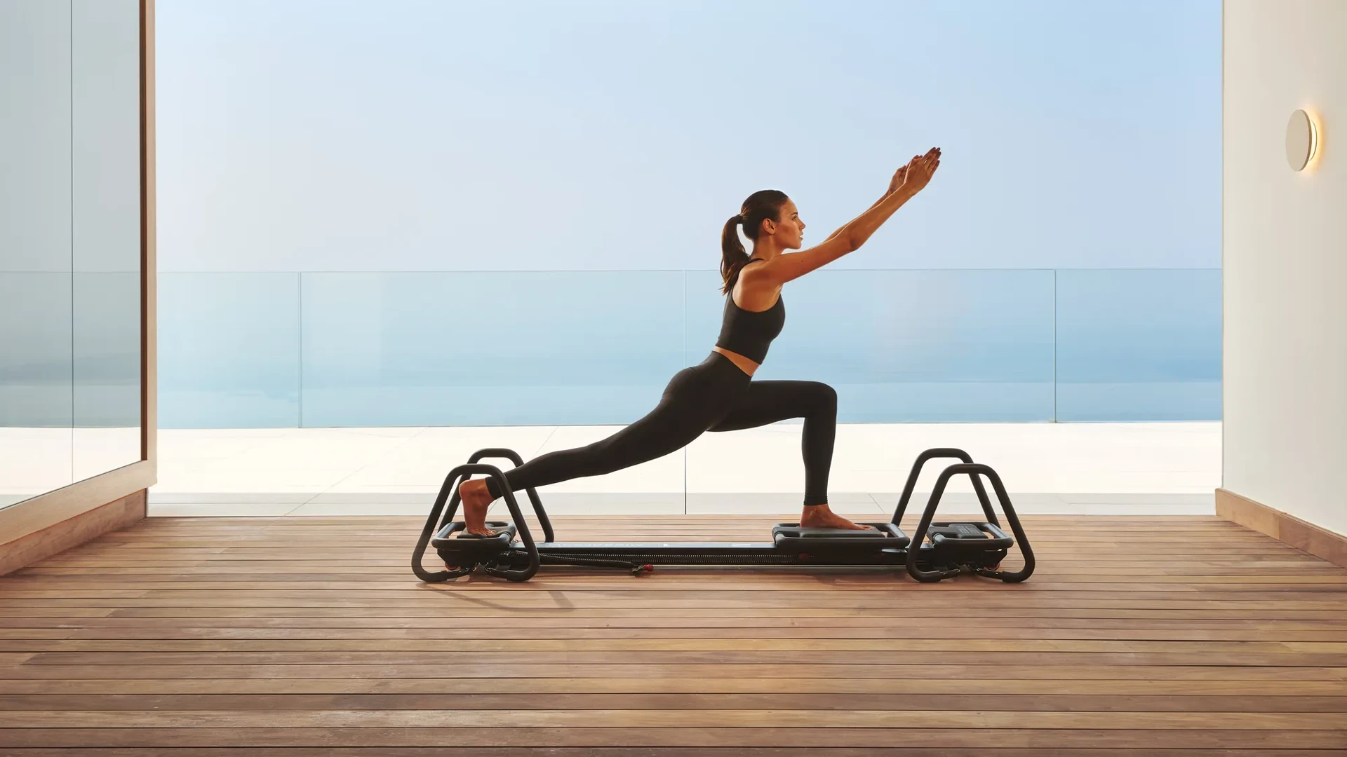 Woman practicing Pilates on a reformer in a modern studio with ocean view through glass walls.