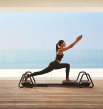 Woman practicing Pilates on a reformer in a modern studio with ocean view through glass walls.