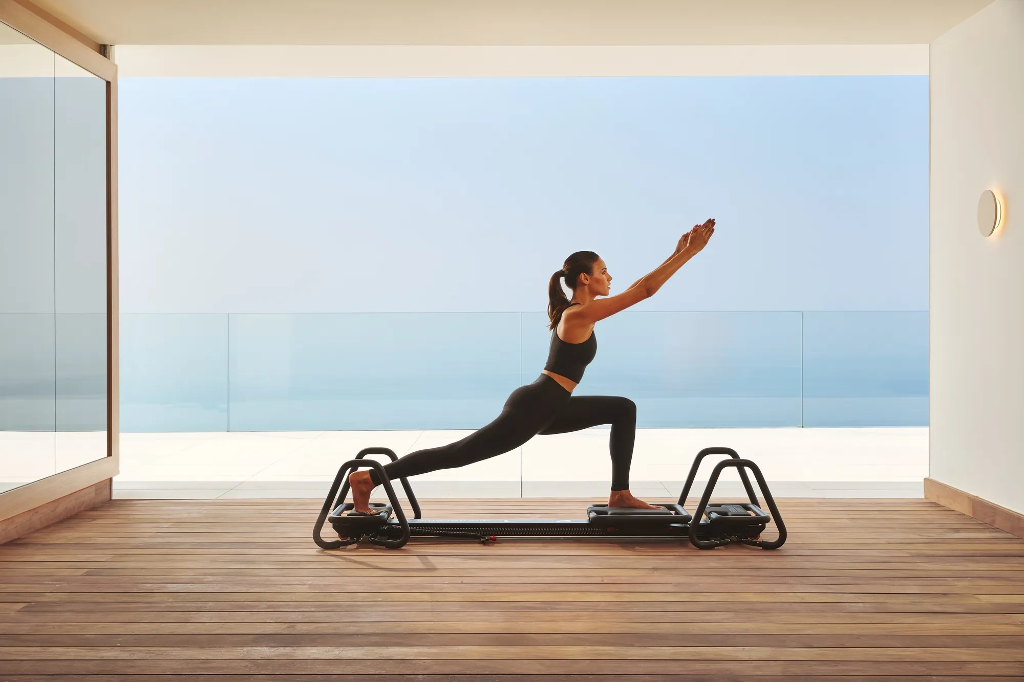 Femme pratiquant le Pilates sur un reformer dans un studio moderne avec vue sur l’océan à travers des vitres.