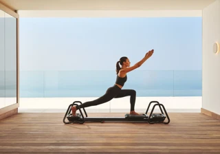 Woman practicing Pilates on a reformer in a modern studio with ocean view through glass walls.