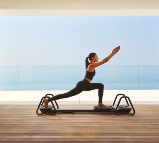 Woman practicing Pilates on a reformer in a modern studio with ocean view through glass walls.