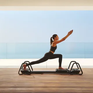 Woman practicing Pilates on a reformer in a modern studio with ocean view through glass walls.