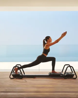 Woman practicing Pilates on a reformer in a modern studio with ocean view through glass walls.