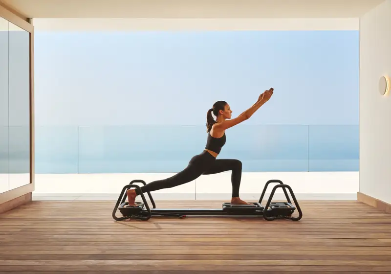 Woman practicing Pilates on a reformer in a modern studio with ocean view through glass walls.