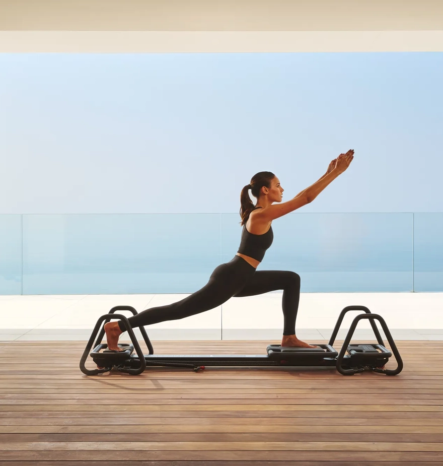 Femme pratiquant le Pilates sur un reformer dans un studio moderne avec vue sur l’océan à travers des vitres.
