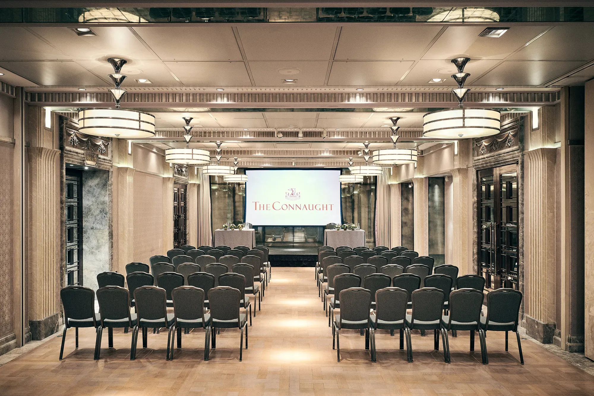 Conference room with rows of chairs facing a screen displaying The Connaught, under warm lighting and elegant detailing.