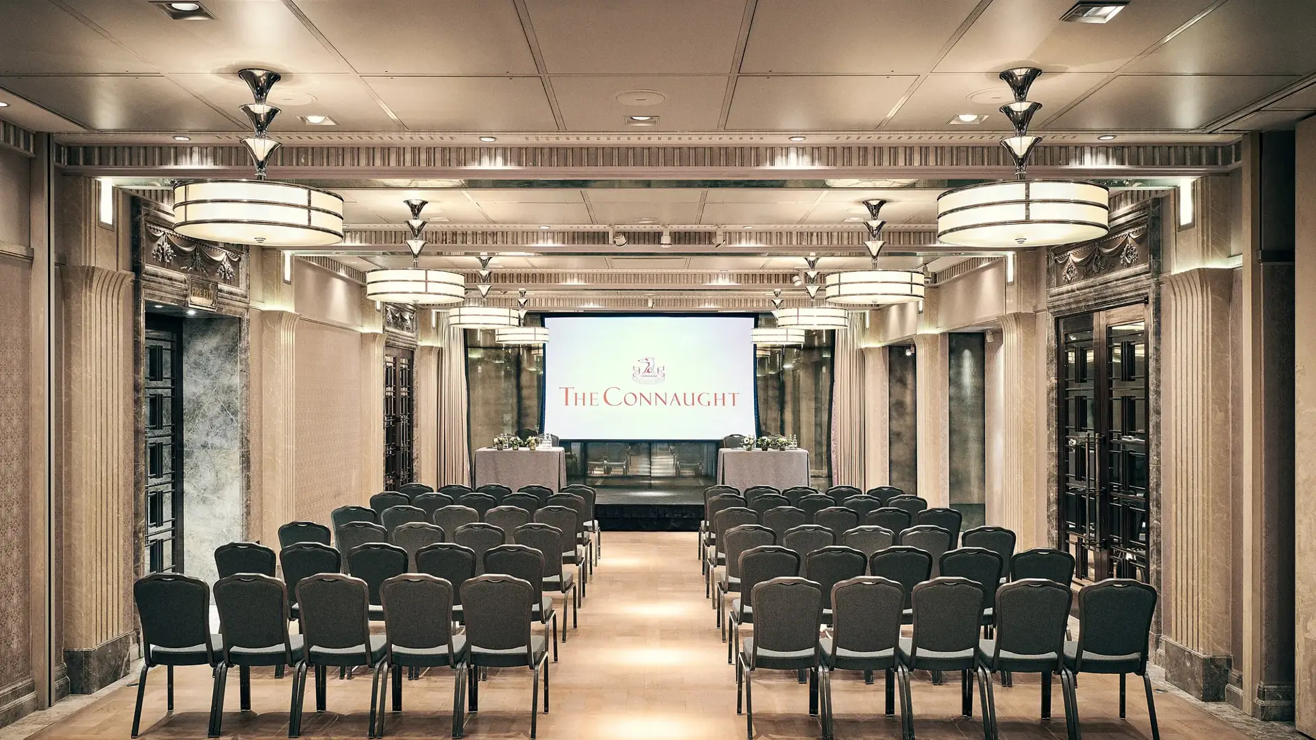 Conference room with rows of chairs facing a screen displaying The Connaught, under warm lighting and elegant detailing.
