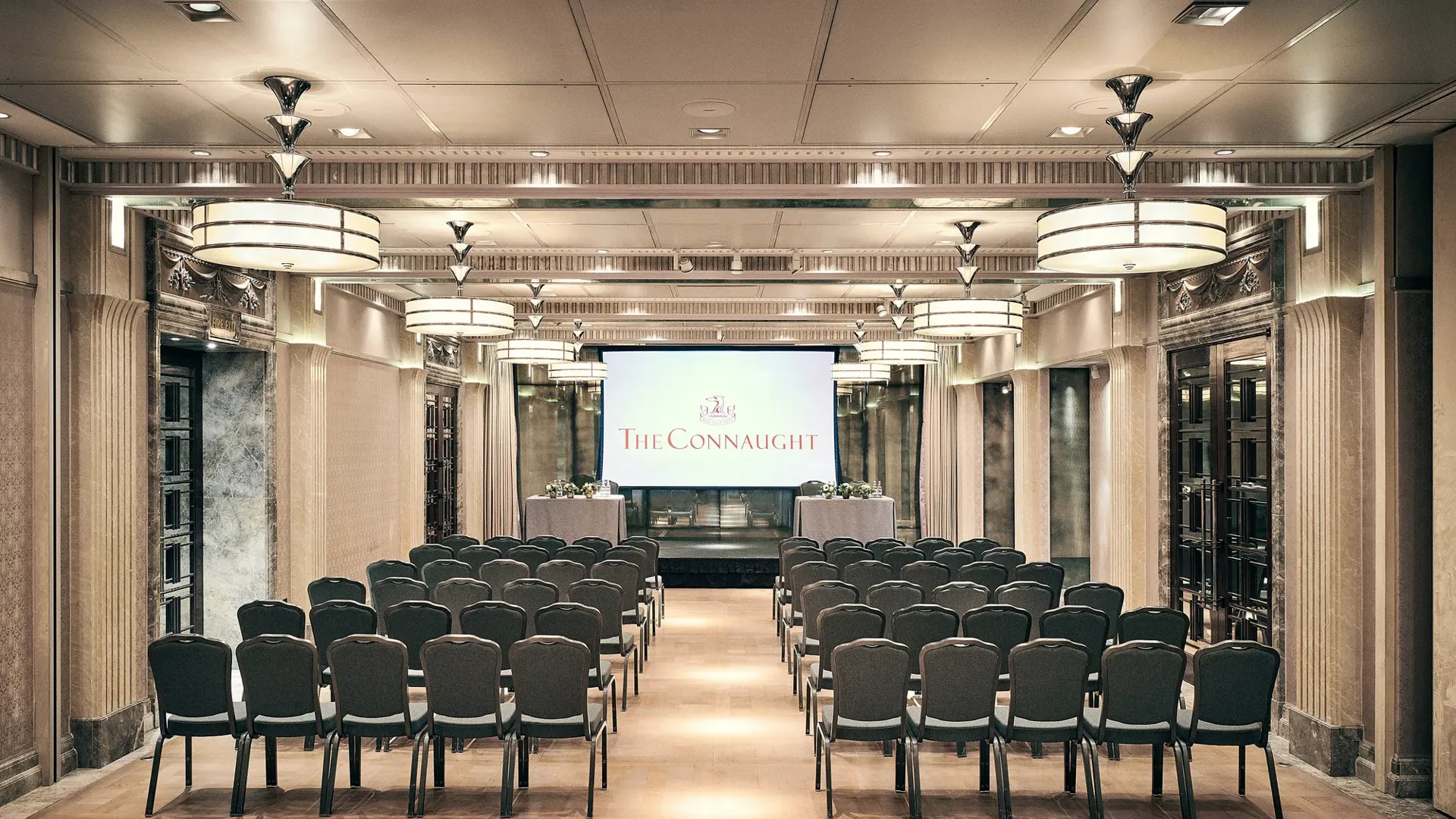 Conference room with rows of chairs facing a screen displaying The Connaught, under warm lighting and elegant detailing.