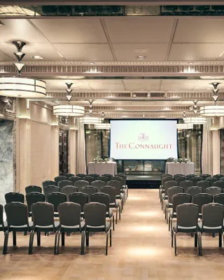 Conference room with rows of chairs facing a screen displaying The Connaught, under warm lighting and elegant detailing.