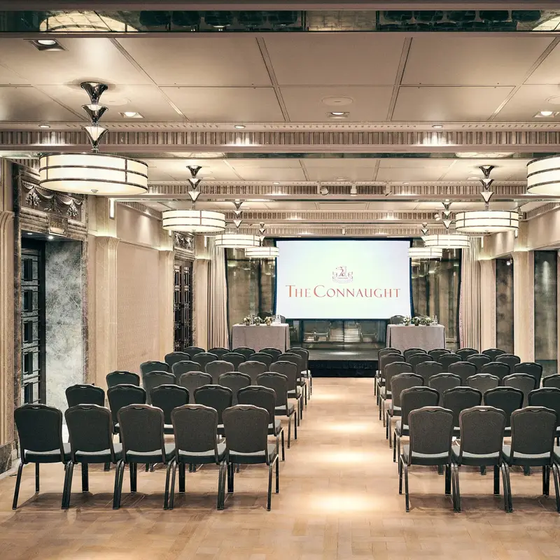 Conference room with rows of chairs facing a screen displaying The Connaught, under warm lighting and elegant detailing.