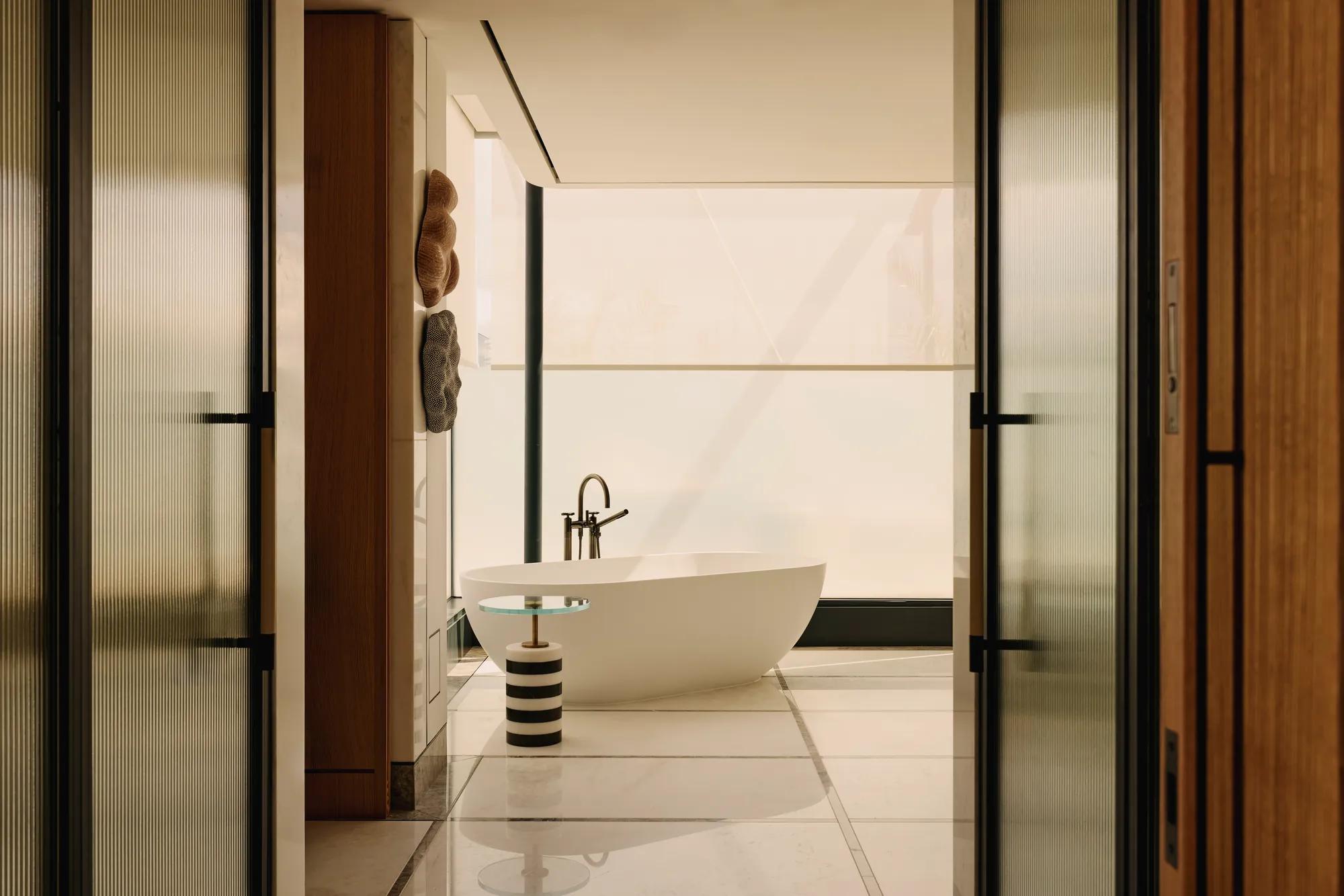 A contemporary freestanding white bathtub is positioned beside a striped side table and sleek black tapware, framed by floor-to-ceiling frosted glass and natural wood panelling in a luxury suite bathroom.