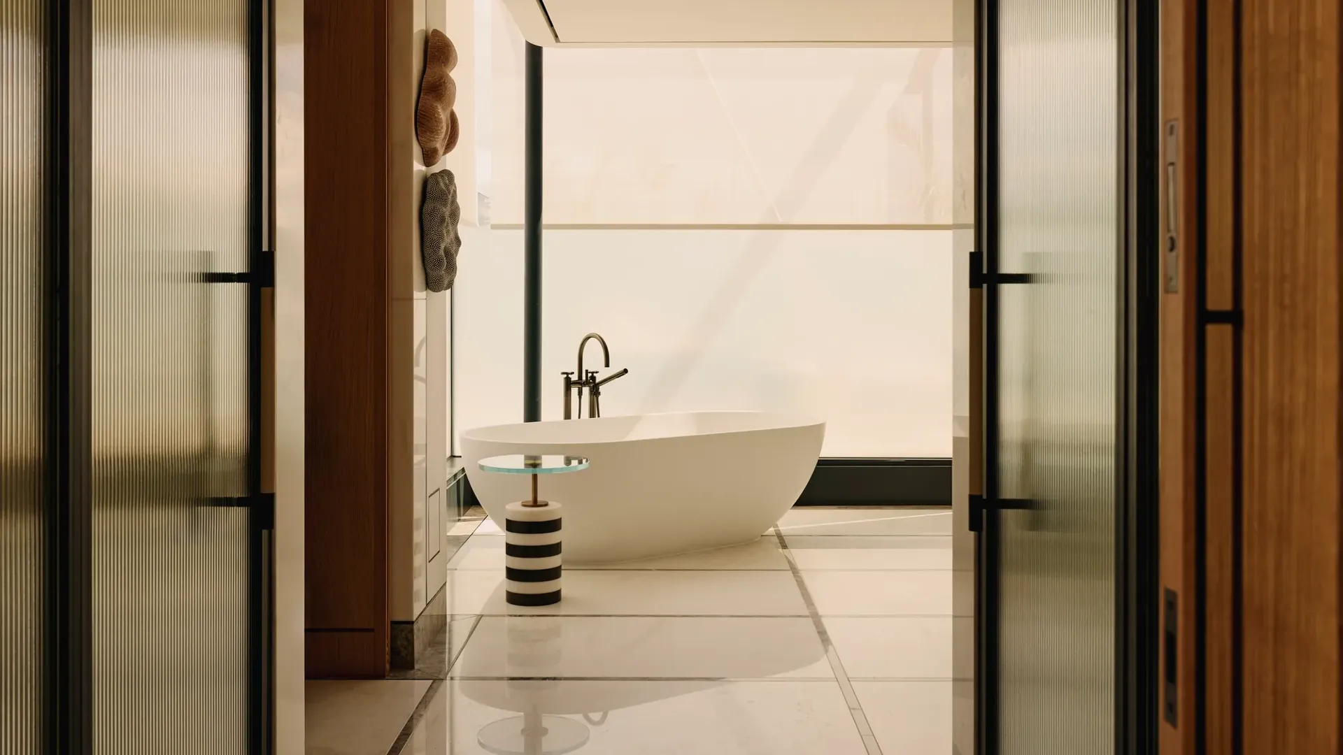 A contemporary freestanding white bathtub is positioned beside a striped side table and sleek black tapware, framed by floor-to-ceiling frosted glass and natural wood panelling in a luxury suite bathroom.