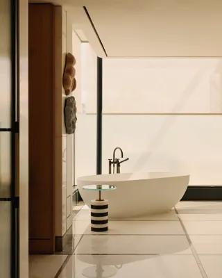 A contemporary freestanding white bathtub is positioned beside a striped side table and sleek black tapware, framed by floor-to-ceiling frosted glass and natural wood panelling in a luxury suite bathroom.