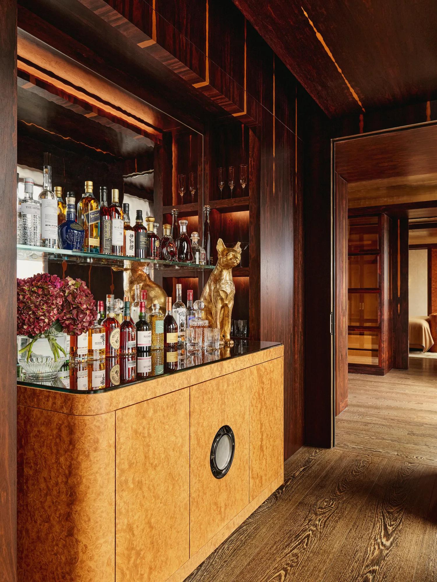 Elegant in-room bar area with a mirrored back display featuring assorted spirits and glassware, a golden dog sculpture, and a light wood cabinet with rounded edges, set against rich dark wood paneling and adjoining a softly lit bedroom.