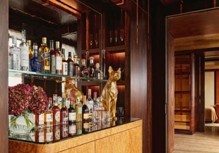 Elegant in-room bar area with a mirrored back display featuring assorted spirits and glassware, a golden dog sculpture, and a light wood cabinet with rounded edges, set against rich dark wood paneling and adjoining a softly lit bedroom.