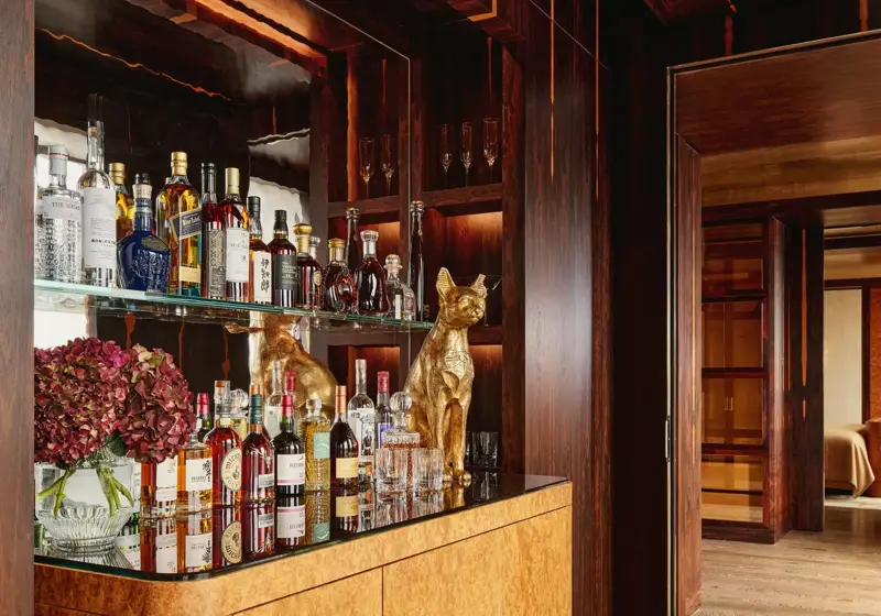 Elegant in-room bar area with a mirrored back display featuring assorted spirits and glassware, a golden dog sculpture, and a light wood cabinet with rounded edges, set against rich dark wood paneling and adjoining a softly lit bedroom.
