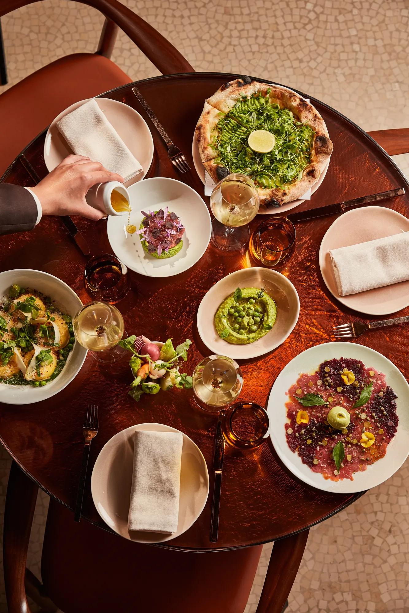 Overhead view of a table filled with green pizza, vibrant starters, wine glasses, and a hand pouring sauce.
