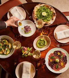 Overhead view of a table filled with green pizza, vibrant starters, wine glasses, and a hand pouring sauce.