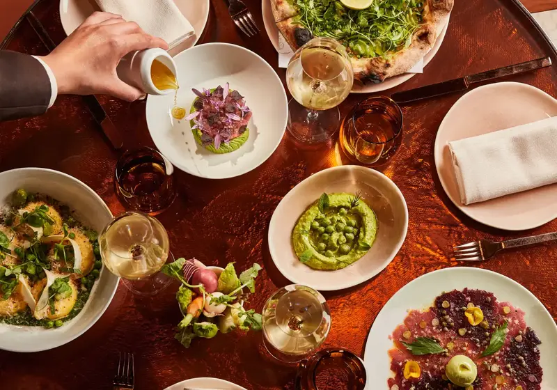 Overhead view of a table filled with green pizza, vibrant starters, wine glasses, and a hand pouring sauce.