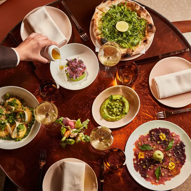 Overhead view of a table filled with green pizza, vibrant starters, wine glasses, and a hand pouring sauce.