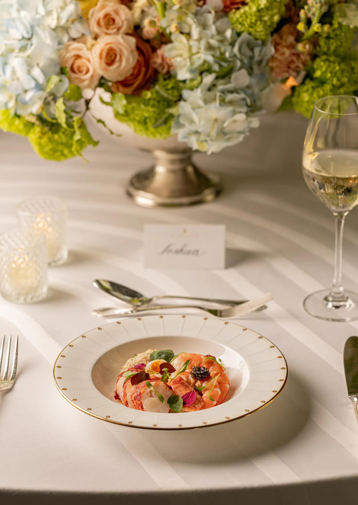 Colourful salmon carpaccio with citrus segments, herbs and edible flowers, presented on fine china at an elegantly dressed table with floral centrepiece and white wine.