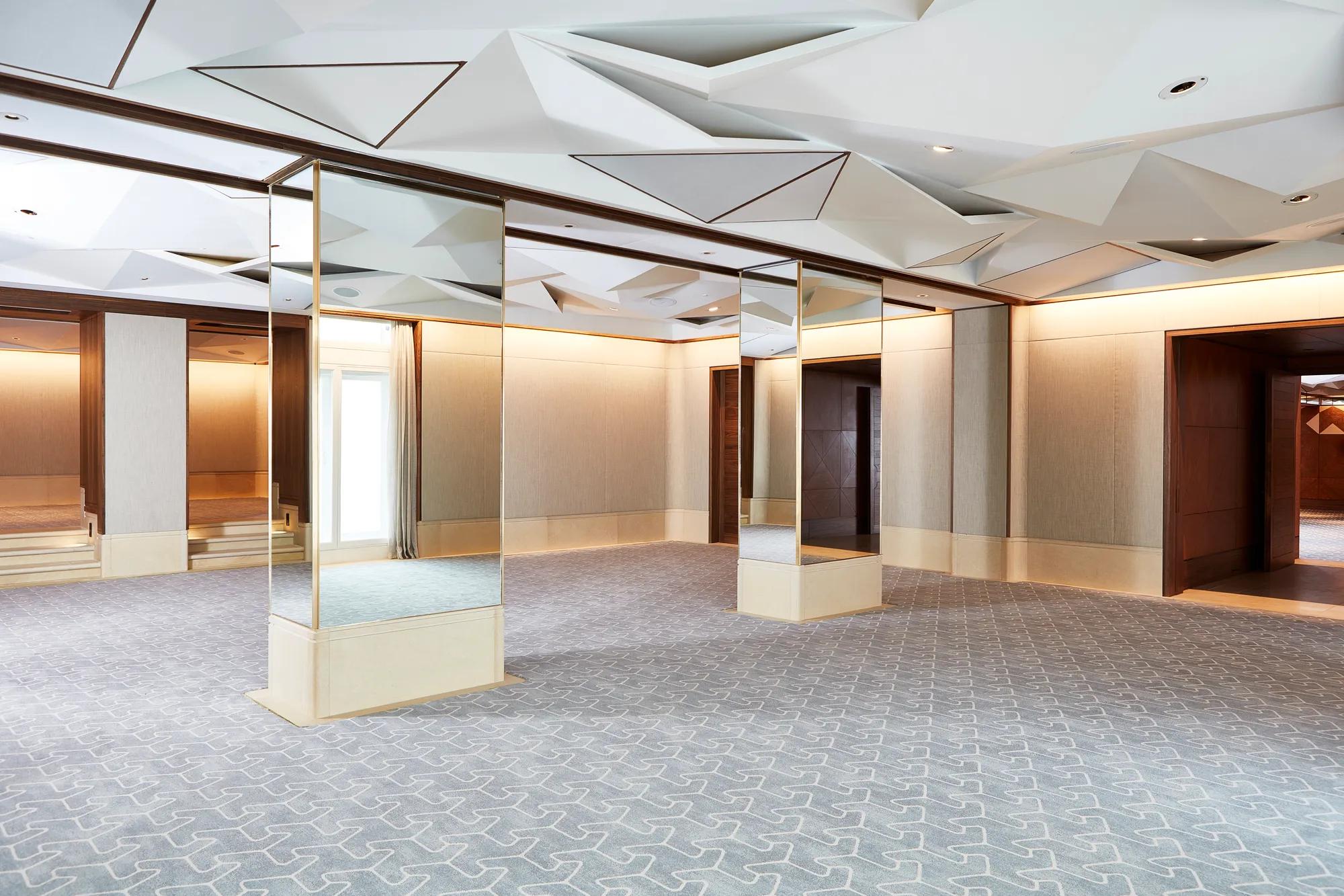 Spacious Belgravia Room with mirrored pillars, geometric ceiling panels, and light grey patterned carpet.