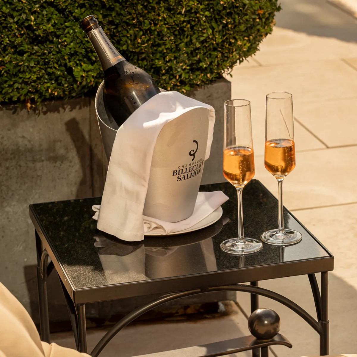 Champagne bottle in an ice bucket with a white cloth and two glasses of rosé champagne on a small outdoor table in the sun.