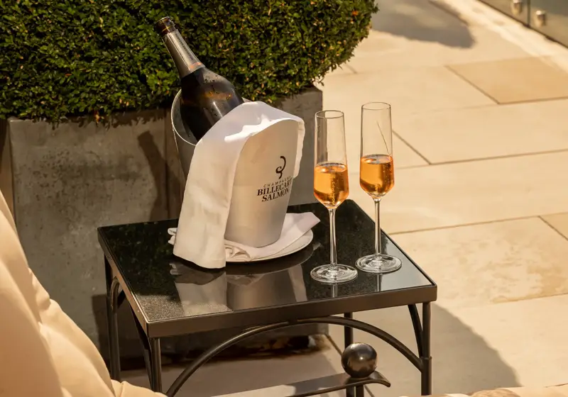 Champagne bottle in an ice bucket with a white cloth and two glasses of rosé champagne on a small outdoor table in the sun.