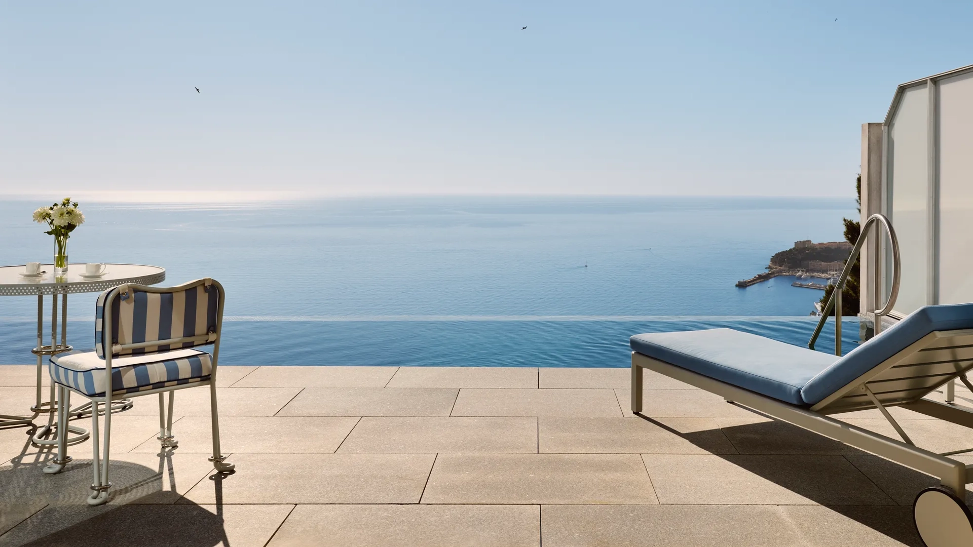 Terrasse avec chaise rayée, table et transat donnant sur une piscine à débordement et une vue panoramique sur la mer sous un ciel bleu clair.