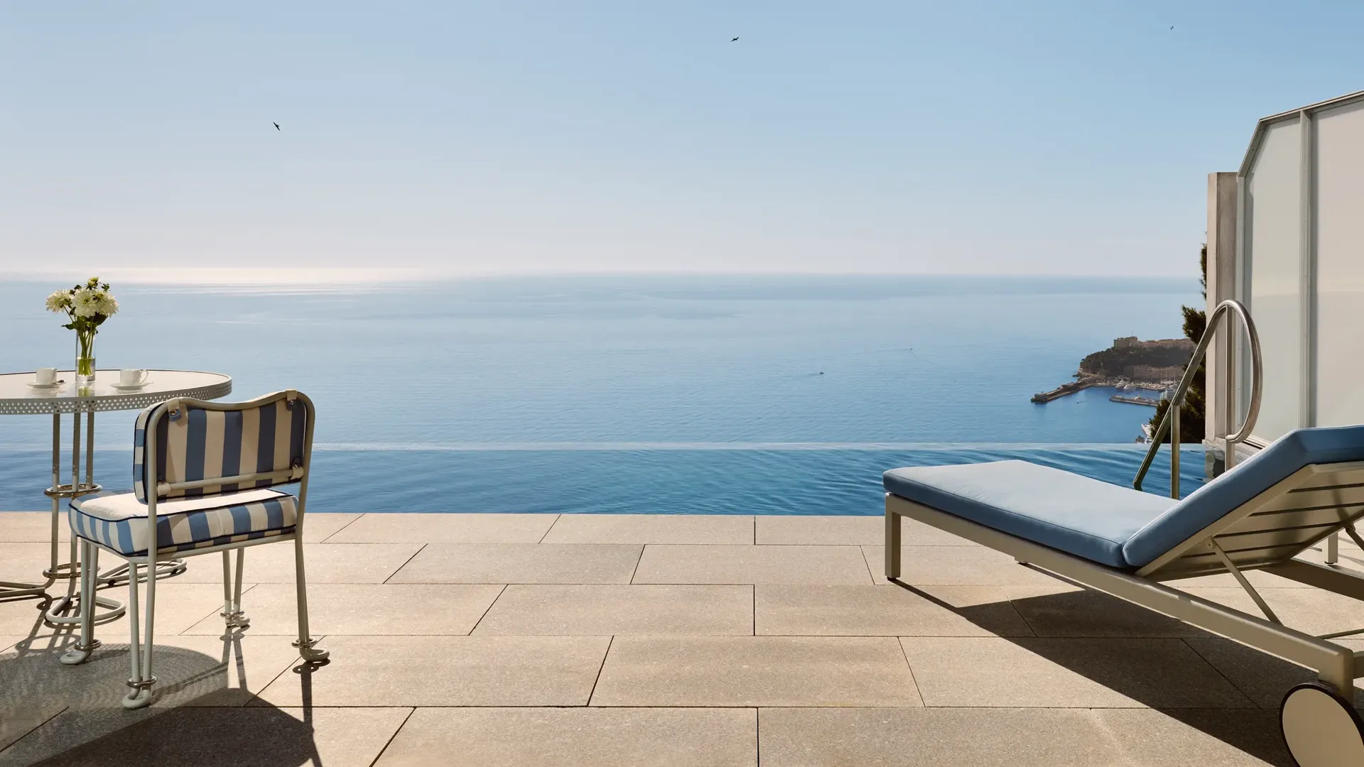 Terrasse avec chaise rayée, table et transat donnant sur une piscine à débordement et une vue panoramique sur la mer sous un ciel bleu clair.