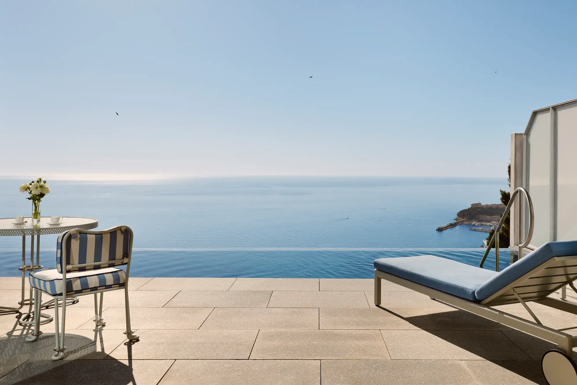 Terrasse avec chaise rayée, table et transat donnant sur une piscine à débordement et une vue panoramique sur la mer sous un ciel bleu clair.
