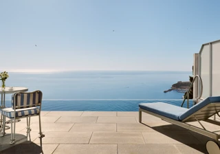 Terrace with striped chair, table, and sun lounger overlooking an infinity pool and panoramic sea view under a clear blue sky.