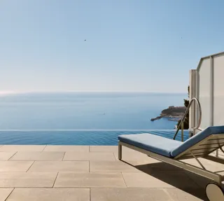 Terrasse avec chaise rayée, table et transat donnant sur une piscine à débordement et une vue panoramique sur la mer sous un ciel bleu clair.