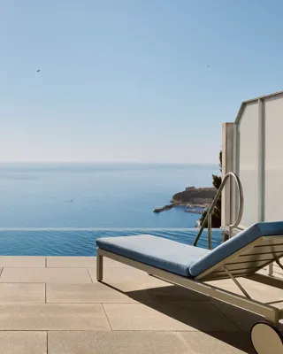Terrasse avec chaise rayée, table et transat donnant sur une piscine à débordement et une vue panoramique sur la mer sous un ciel bleu clair.