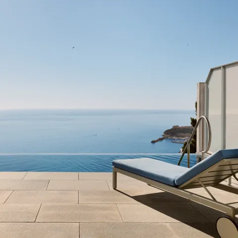 Terrasse avec chaise rayée, table et transat donnant sur une piscine à débordement et une vue panoramique sur la mer sous un ciel bleu clair.